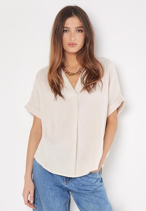 Blouse beige clair à manches courtes, avec un décolleté en V et une finition texturée, associée à un jean bleu ample. Présente une coupe décontractée et des accents subtils.