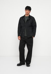 Black jacket with front pockets, buttoned closure, and khaki accents, worn over a dark green sweater and paired with loose black trousers.