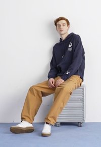 Hanorac bleumarin cu logo, pantaloni largi bej și adidași albi cu talpă maro, așezat pe un scaun metalic cu striații, în fața unui perete deschis.