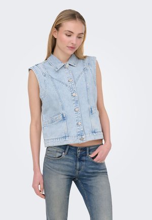 Tânără purtând o vestă de denim albastru deschis fără mâneci cu nasturi și blugi albaștri, stând în fața unui fundal simplu și deschis.