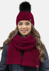 Maroon knitted beanie and scarf set featuring a textured pattern, with a black faux fur pom-pom on the hat. Soft, warm texture.