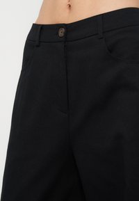 Gros plan d’un pantalon noir à fermeture boutonnée porté par une personne, montrant la ceinture, les passants et le ventre partiellement nu avec le nombril visible.