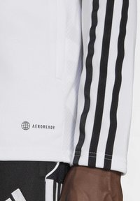 Witte sportshirt met zwarte strepen langs de mouwen, textuurstof en een "AEROREADY" logo op de zijkant.