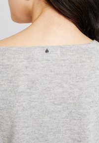Grauer Strickpullover mit weitem Ausschnitt, ausgestattet mit einem kleinen dunklen Metallelement im Rückenbereich. Weiche Textur mit einem dezenten Muster.