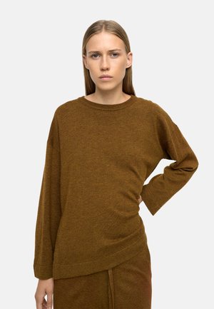 Młoda kobieta z prostymi blond włosami ubrana w brązowy sweter z długim rękawem i dopasowane spodnie, stojąca z jedną ręką na biodrze.