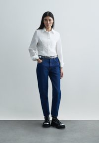 Balti marškiniai ilgomis rankovėmis, derinami su mėlynais aukšto liemens džinsais ir juodais bateliais su raišteliais ir blizgiomis koja apdailomis.