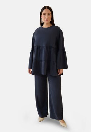 Kvinde med langt mørkt hår iført navyblå lagdelt bluse og bukser med vide ben, beige spidse sko og store statement-øreringe.