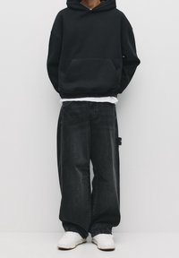 Černá mikina s kapucí a přední kapsou, doplněná volnými tmavě šedými džínami s opranými detaily.