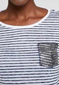Camisa de rayas en blanco y azul marino, con un cuello redondo y un bolsillo en el lado izquierdo del pecho adornado con lentejuelas. Textura de tela suave.