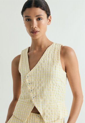 Mujer con chaleco de cuadros vichy amarillo y blanco sin mangas con cuatro botones sobre pantalones a juego, de pie sobre un fondo liso.