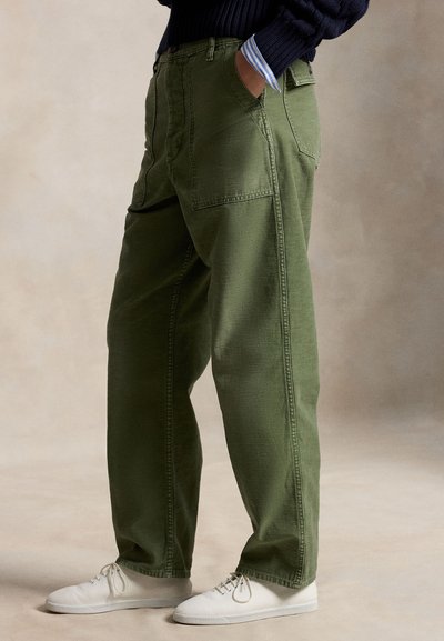 Persoană purtând pantaloni largi verde măsliniu cu buzunare în față și în spate, pulover bleumarin, manșetă de cămașă cu dungi și pantofi albi cu șireturi.