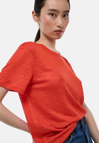 T-shirt rouge à manches courtes avec un col en V, fabriqué en tissu léger, présentant une texture subtile et une coupe décontractée. Porté avec un jean taille haute.