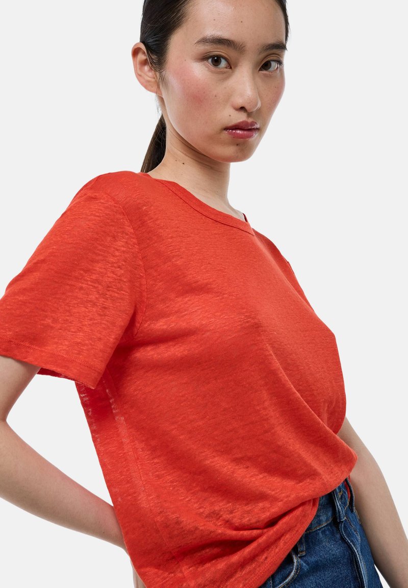 T-shirt rouge à manches courtes avec un col en V, fabriqué en tissu léger, présentant une texture subtile et une coupe décontractée. Porté avec un jean taille haute.