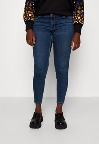 Dark blue skinny jeans with a smooth texture, paired with black loafers. The jeans feature a classic five-pocket design and a button closure.