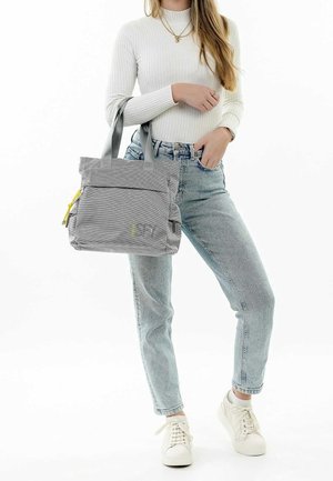 Grå tote-bag i tekstureret stof, med to håndtag, gul lynlåsdetalje og frontlomme, vist mens den holdes med et afslappet outfit.