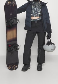 Snowboard avec des fixations noires, une doudoune sombre, un haut à motifs, un pantalon noir et un casque gris. Caractérise des matériaux durables et un design fonctionnel.