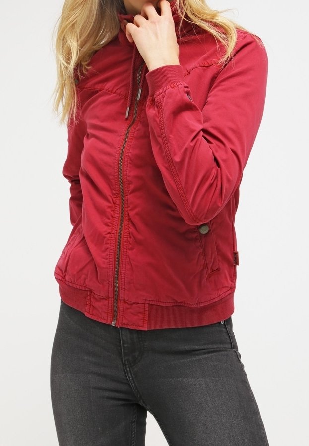 Femme portant une veste rouge ajustée zippée et un jean gris foncé, une main près du visage et l’autre dans la poche.