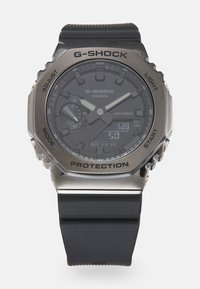 Montre G-SHOCK avec un bracelet en caoutchouc noir et un cadran foncé, dotée d'accents en argent, d'un affichage numérique et d'une lunette en métal protectrice.