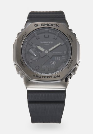 Zegarek G-SHOCK na nadgarstek z czarnym gumowym paskiem i ciemną tarczą, z metalicznymi srebrnymi akcentami, wyświetlaczem cyfrowym oraz ochronnym metalowym pierścieniem.