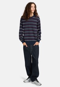 Maglione a righe navy in lavorazione a maglia con righe multicolori, polsini e orlo a costine, abbinato a pantaloni in jeans scuri e scarpe sportive argentate.