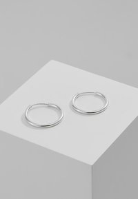 Pendientes de aro de plata con un diseño circular y suave. Perfil delgado y acabado pulido sobre un pedestal gris claro. Sencillos y elegantes.