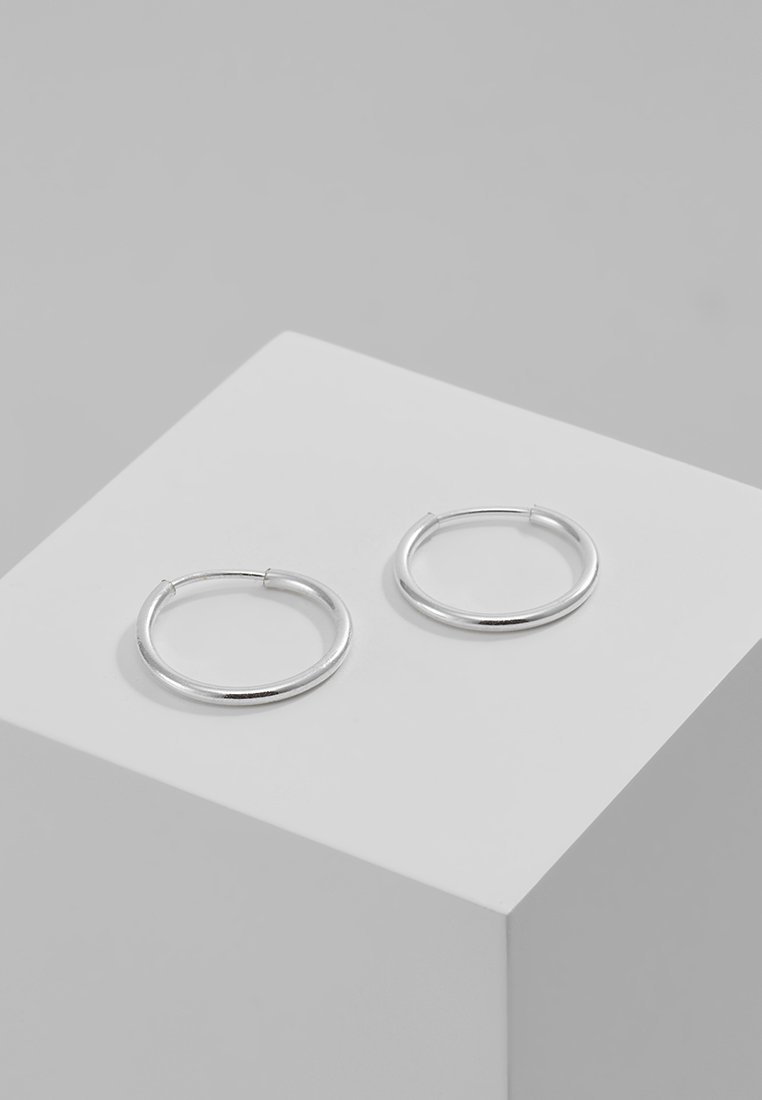 Pendientes de aro de plata con un diseño circular y suave. Perfil delgado y acabado pulido sobre un pedestal gris claro. Sencillos y elegantes.
