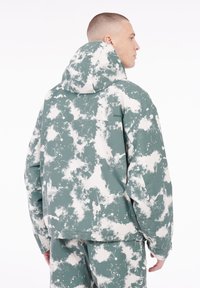 Kapuzenshirt mit einem grau-weißen Wolkenmuster. Hergestellt aus glattem Stoff, bietet eine lockere Passform und eine Brusttasche mit Reißverschluss.