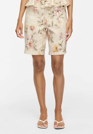 Femme portant un short beige à fleurs jusqu'aux genoux et des sandales blanches, debout devant un fond blanc uni.