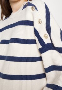 Suéter crema con rayas horizontales en navy. Presenta textura acanalada y acentos de botones en tono dorado en el hombro. Corte relajado.