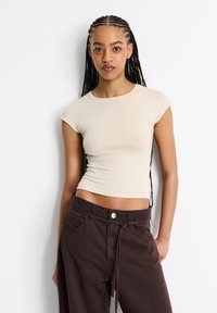 Beiger Baumwoll-Crop-Top mit kurzen Ärmeln, kombiniert mit dunkelbraunen, hochgeschnittenen Hosen mit Knopfverschluss und Bindeband.
