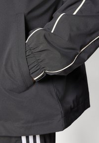 Veste noire avec des poignets élastiques, accents de passepoil blanc et poches latérales. Le matériau semble lisse avec une finition légèrement mate.