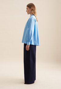 Hellblauses, oversized Hemd mit langen Ärmeln und Knopfmanschetten, kombiniert mit hochtaillierten, navyblauen, weiten Hosen, vor einem neutralen Hintergrund.