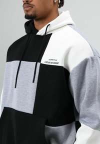 Color-blocked hoodie in black, white, and gray. Features a kangaroo pocket and embroidered text “LIVE IN THE MOMENT.” Drawstring hood.