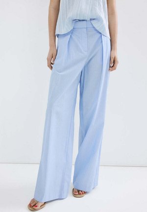 Femme portant un pantalon large bleu clair à rayures verticales et des sandales beiges, debout devant un fond blanc uni.