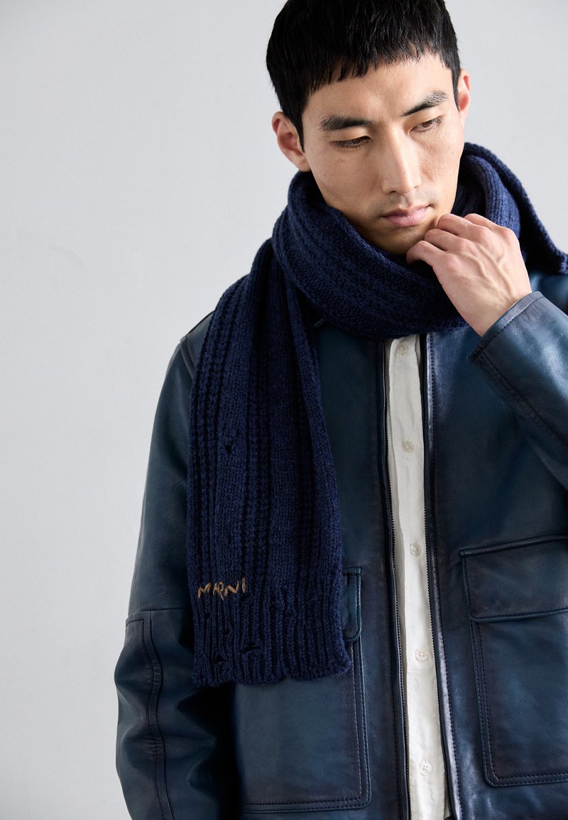 Een jonge man die een marineblauwe gebreide sjaal over een wit shirt en een donkere leren jas aanpast, terwijl hij naar beneden kijkt tegen een eenvoudige achtergrond.