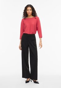 Vrouw met krullend haar, gekleed in een rood shirt met lange mouwen, een zwarte hooggesneden wijde broek en zwarte puntige hakken, staand voor een witte achtergrond.