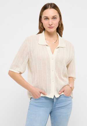 Woman with light brown hair wearing gold hoop earrings, a cream short-sleeve textured button-up shirt, light blue jeans, and gold jewelry with hands in pockets.