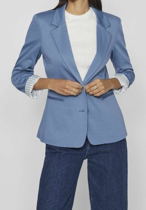 Kobieta zapinająca jasnoniebieską marynarkę z podwiniętymi, paskowanymi mankietami, noszoną na białej bluzce i ciemnoniebieskich dżinsach, na jednolitym tle.