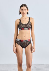 Μαύρο σετ εσωρούχων από δαντέλα με σουτιέν που διαθέτει φαρδιά λωρίδα λογότυπου σε κόκκινο και μαύρο χρώμα και αντίστοιχα σλιπ, αναδεικνύοντας περίπλοκα σχέδια και υφές.