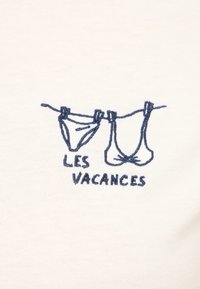 Tela blanca con bordados azules de dos trajes de baño colgados en un tendedero, con el texto "LES VACANCES" debajo.
