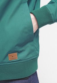 Grøn jakke med en glat tekstur, ribbede manchetter og kant, med et læderlogo patch mærket "TWIN TIP" i taljen.