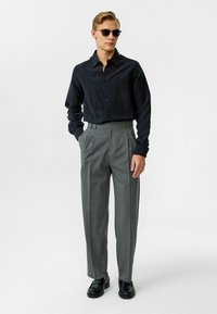 Zwarte overhemd met knopen, grijs geplooide broek en zwarte schoenen. Glad fabric met een subtiele textuur, perfecte pasvorm en zonnebril.