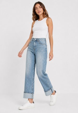 Vrouw met een mouwloos wit topje, hoog getailleerde, wijde blauwe jeans met omslagen en witte sneakers, staand tegen een effen achtergrond.