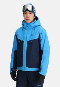 Blauwe en marineblauwe skijas met twee ritsborstzakken, verstelbare capuchon en zwarte handschoenen. Ontworpen voor koud weer met ventilatiefuncties.