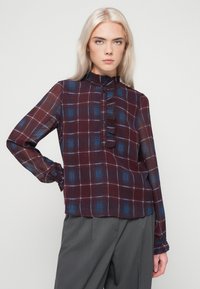 Fiatal nő, aki szőke hajú, bordó és kék kockás blúzt és szürke nadrágot visel, egyik kezét a csípőjén támasztva áll.