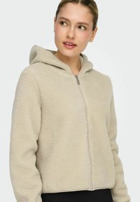 Beige fleece-hettejakke med teksturert overflate, frontglidelås og hette. Designet har en lett kortet passform og avslappede ermer.
