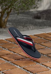 Flip-flop navy cu bretele cu dungi roșii, albe și negre și logo-ul Tommy Hilfiger, plutind deasupra unei pardoseli exterioare din plăci de lemn.