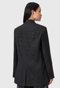 Donna con capelli lunghi e scuri che indossa un blazer nero con testo forato che recita "ALL SAINTS" sulla schiena, in piedi contro uno sfondo semplice.