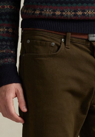 Brune bomullsbukser med en jevn tekstur, utstyrt med metallbeslag, sidelommer og et brunt lærbelte, delvis skjult av en mønstret strikkegenser.