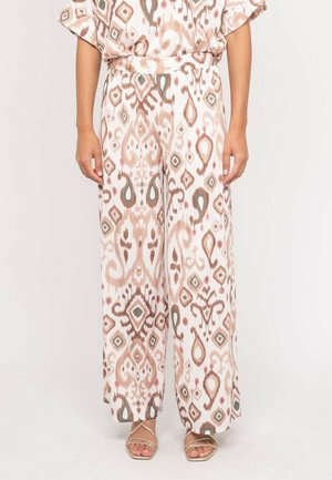 Femme portant un pantalon à jambes larges avec des motifs abstraits beiges, marron et roses, assorti à des sandales beige à brides.
